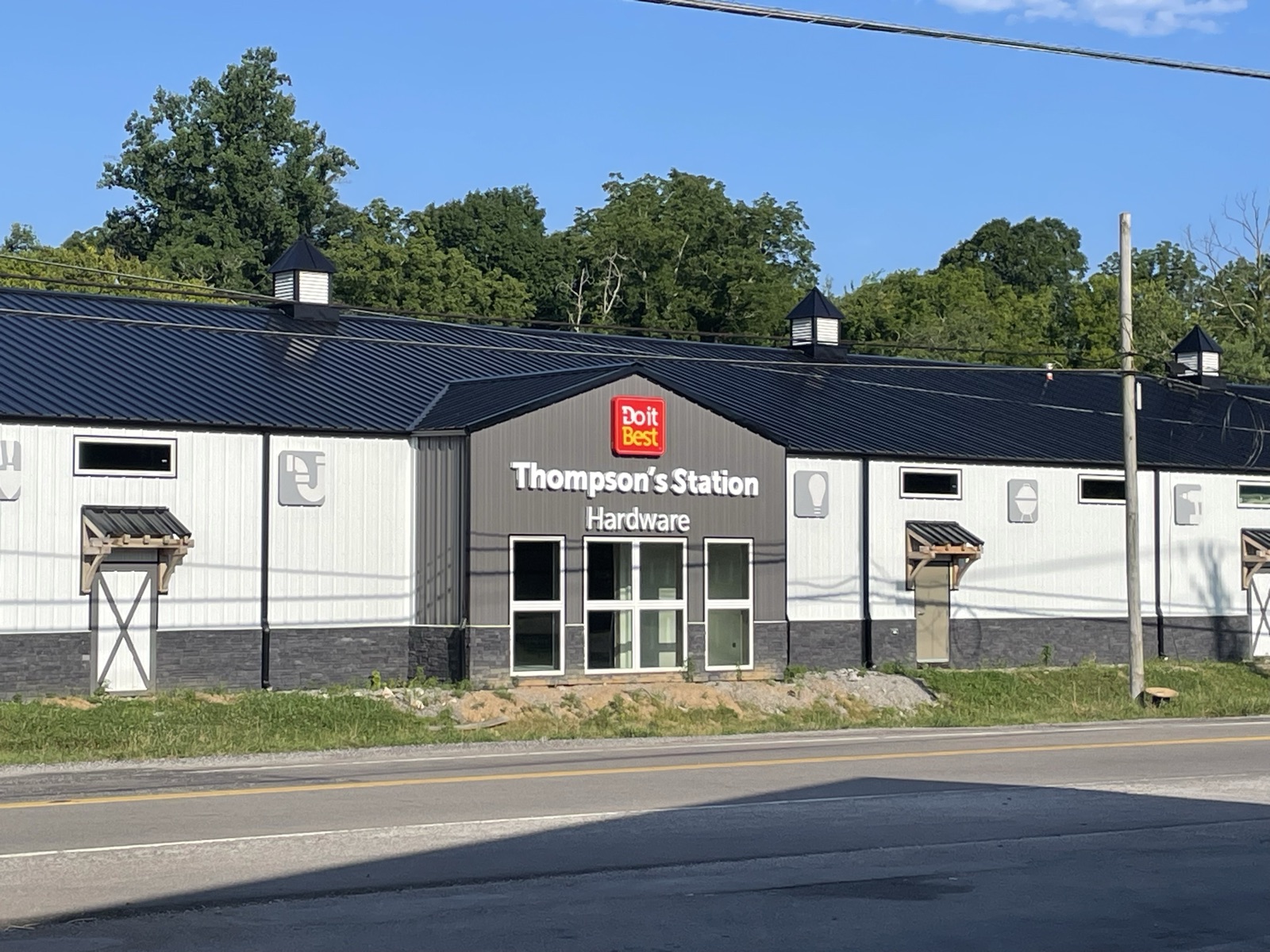 Completed Thompson's Station Hardware storefront viewed from across Lewisburg Pike, with the illuminated Do it Best sign and store name mounted on the central gable and finished white metal siding.